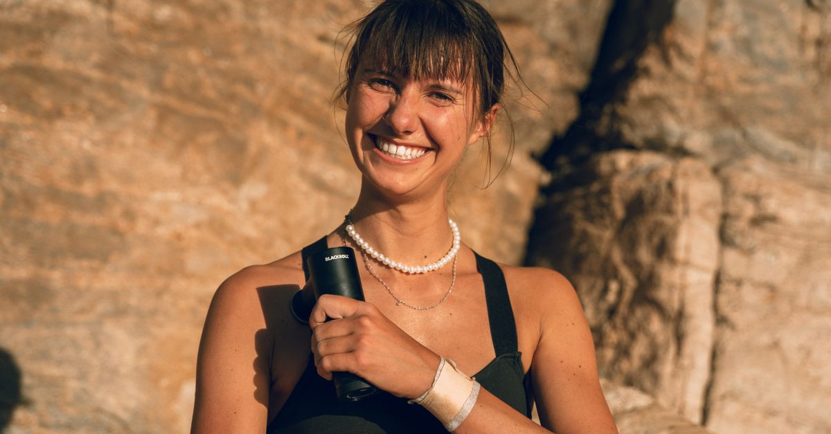 A woman smiles while holding a massage gun in a sunny, rocky outdoor setting.